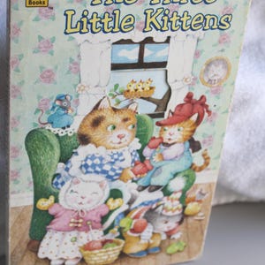 May include: A vintage children's book titled "The Three Little Kittens" with a colorful illustration. The cover features three anthropomorphic kittens, a mouse, and a bird's nest, set against a floral wallpaper background. The Golden Books logo is visible.