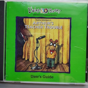 May include: A Living Books CD case for Marc Brown's Arthur's Teacher Trouble. The green cover features an illustration of Arthur standing on a stage with a microphone, while another character peeks from behind a yellow curtain. Text reads User's Guide.