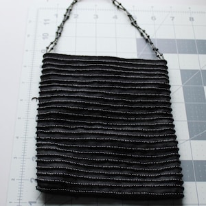 May include: A black beaded handbag with a beaded handle. The rectangular bag features horizontal rows of black fabric with small, white beads. The handle is made of small, black beads. The bag is on a white surface with a grid pattern.