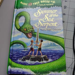 May include: A hardcover book titled Magic Tree House #31 A Merlin Mission Summer of the Sea Serpent by Mary Pope Osborne. The cover features a large green sea serpent encircling two children standing on a rock in the ocean holding a glowing sword.