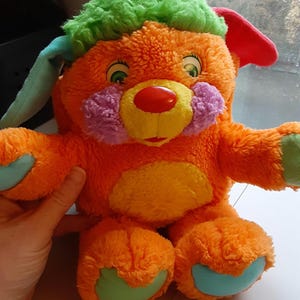 May include: An orange plush toy with green hair, blue and red ears, and a red nose. The toy has purple cheeks, a yellow mouth, and blue accents on its feet and hands. It is a colorful, whimsical stuffed animal.