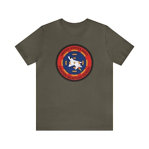 May include: A dark green t-shirt with a circular patch featuring a fighter jet and the text "United States Navy Fighter Weapons School".