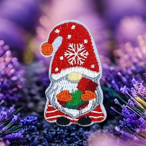 Puede incluir: Parche bordado de gnomo navideño con gorro rojo, barba blanca y diseño de copo de nieve. El gnomo sostiene una corona verde y naranja. El parche tiene un borde blanco sobre un fondo morado borroso.