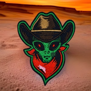 May include: Green alien wearing a black cowboy hat with a red bandana. The alien has large green eyes and a green face. The image is a patch.