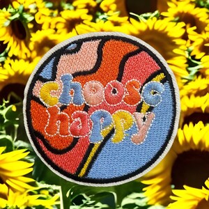 May include: A colorful embroidered patch with a swirling design and the text "choose happy" in a retro font. The patch is surrounded by a field of yellow sunflowers.