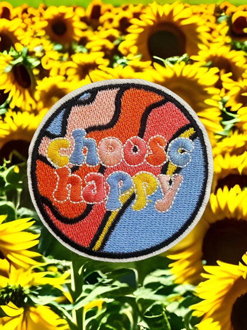 May include: A colorful embroidered patch with a swirling design and the text "choose happy" in a retro font. The patch is surrounded by a field of yellow sunflowers.