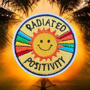 May include: A yellow embroidered patch with a smiling sun and rainbow rays. The text "RADIATE POSITIVITY" is stitched around the sun in black thread.