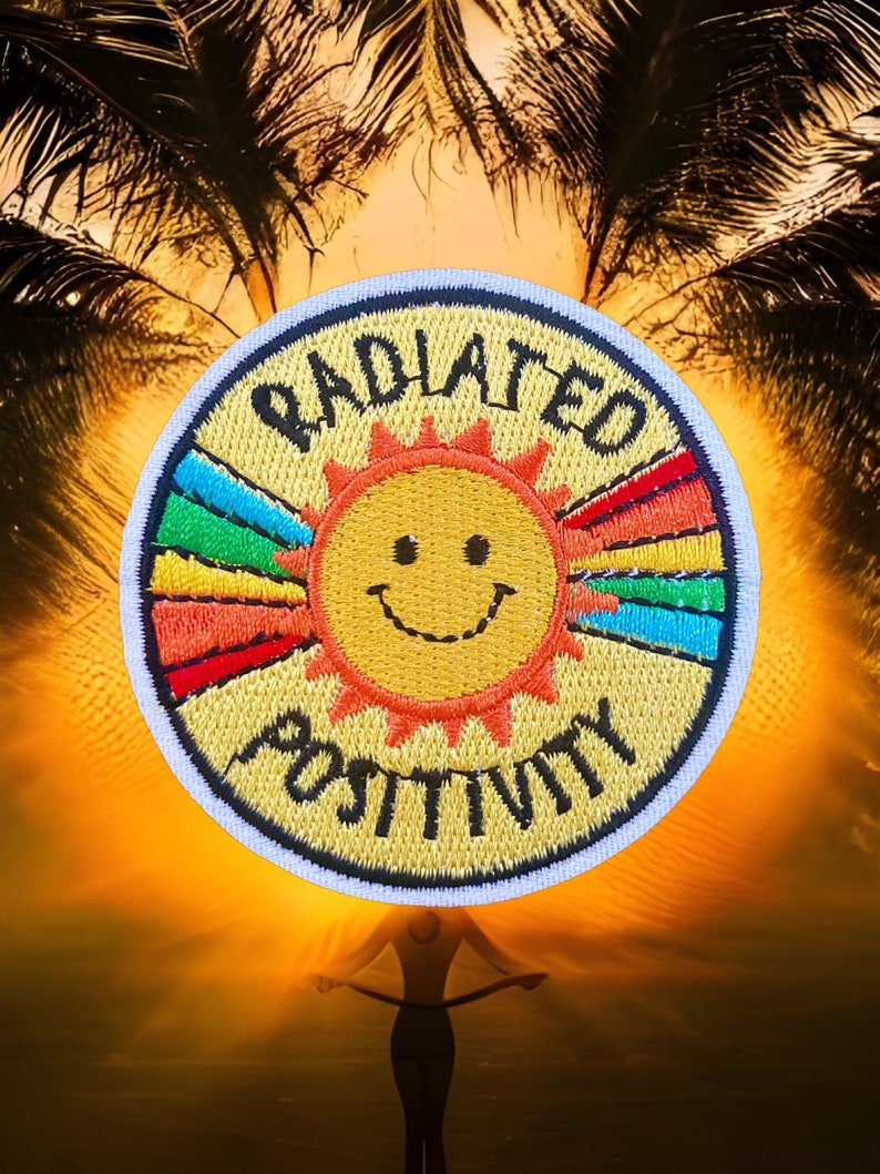 May include: A yellow embroidered patch with a smiling sun and rainbow rays. The text "RADIATE POSITIVITY" is stitched around the sun in black thread.