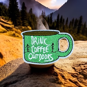 May include: Green embroidered patch of a coffee mug with the text "Drink Coffee Outdoors" on a wooden surface with a blurry background of a mountain range.