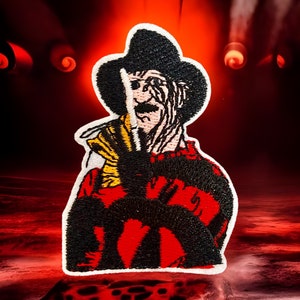 Könnte beinhalten: Ein gestickter Aufnäher mit einem Horrorfilmcharakter. Der Charakter trägt einen Hut und ein gestreiftes Hemd. Der Charakter hat ein weißes Gesicht mit einem schwarzen Auge und einem schwarzen Mund.