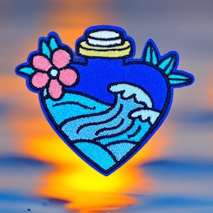 Puede incluir: Parche bordado en forma de corazón, con un diseño de botella azul con una flor rosa y olas del océano. El parche tiene un borde azul y está sobre un fondo borroso de puesta de sol.