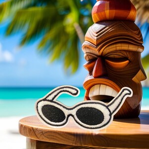 May include: White sunglasses with a black lens, sitting on a wooden table in front of a carved wooden tiki statue. The statue is brown and red with a straw-like top.