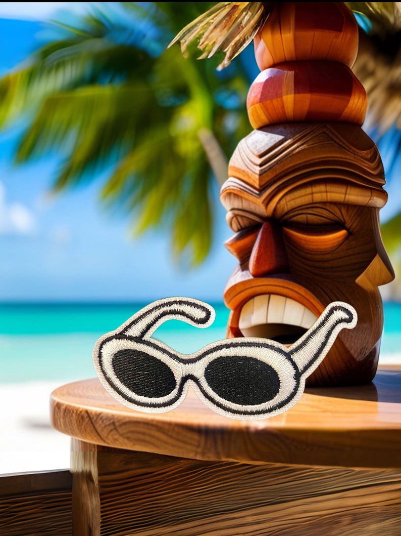 May include: White sunglasses with a black lens, sitting on a wooden table in front of a carved wooden tiki statue. The statue is brown and red with a straw-like top.