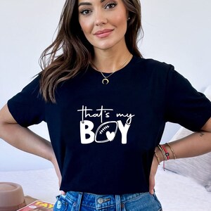 May include: Black t-shirt with white text that reads "That's my BOY" with a football and heart graphic.