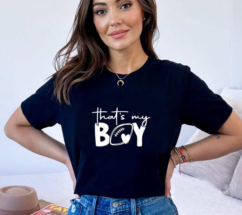 May include: Black t-shirt with white text that reads "That's my BOY" with a football and heart graphic.