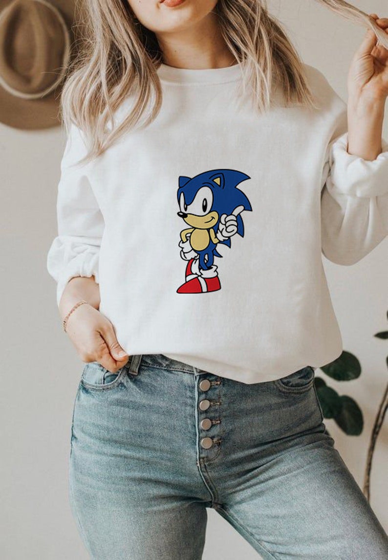 May include: White sweatshirt with a blue and yellow cartoon character wearing red shoes. The character is giving a thumbs up. The sweatshirt is being worn by a person with long blonde hair.