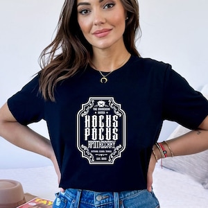 May include: A black t-shirt featuring a white graphic of a vintage label that reads "The Sanderson Sister + Hocus Pocus Apothecary, Potion Elixir Things, Est. 1933."