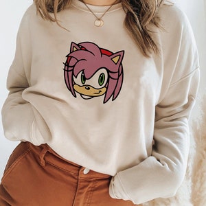 May include: A cream-colored sweatshirt with a pink and green cartoon character on the front. The character has pink hair, green eyes, and a yellow nose.