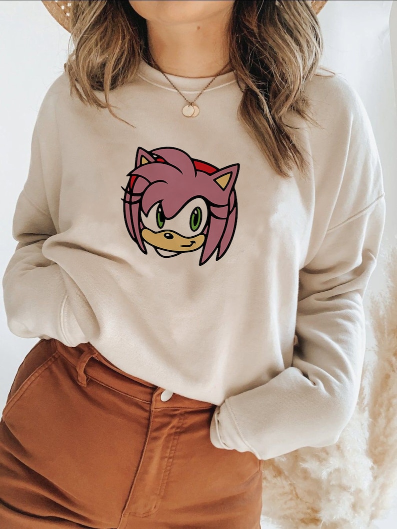 May include: A cream-colored sweatshirt with a pink and green cartoon character on the front. The character has pink hair, green eyes, and a yellow nose.