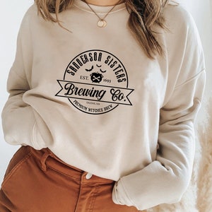 May include: A beige sweatshirt with a black graphic print that reads "Sanderson Sisters Brewing Co. Est. 1693 Salem, MA Premium Witches Brew".