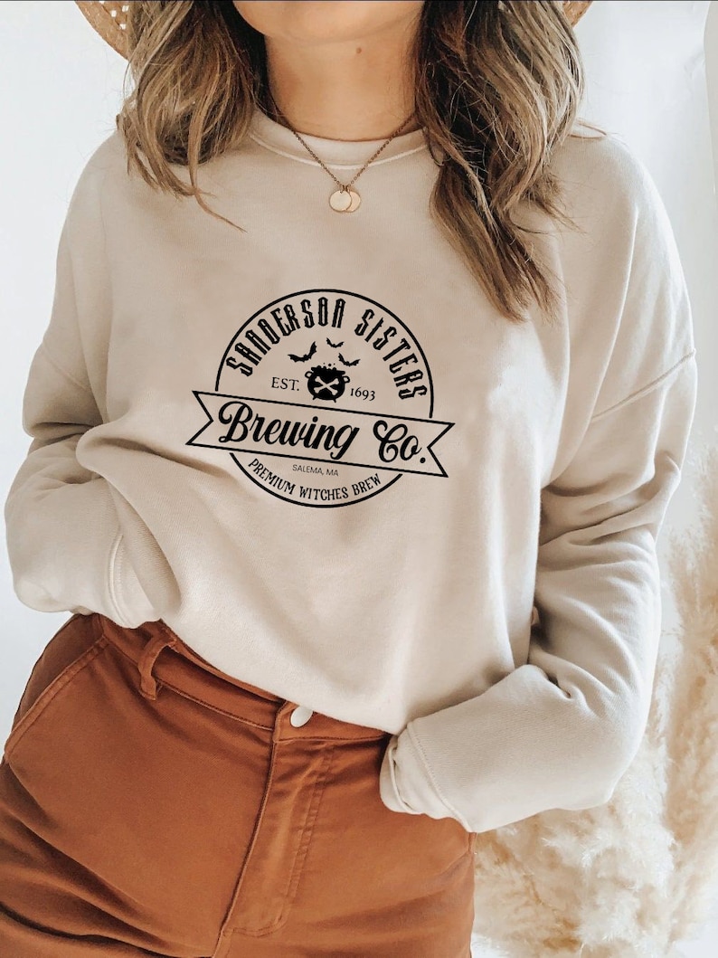 May include: A beige sweatshirt with a black graphic print that reads "Sanderson Sisters Brewing Co. Est. 1693 Salem, MA Premium Witches Brew".