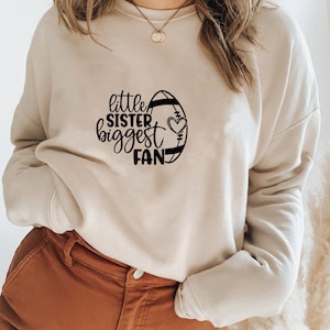May include: A beige sweatshirt with a black graphic design of a football with the text "little sister biggest fan" and a heart.