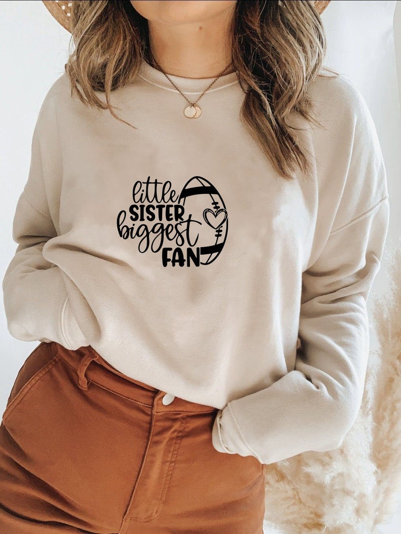May include: A beige sweatshirt with a black graphic design of a football with the text "little sister biggest fan" and a heart.