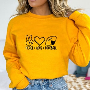 May include: Yellow sweatshirt with a peace sign, heart, and football graphic. The text reads "Peace Love Football".
