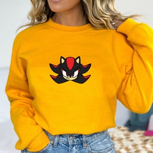 May include: A yellow sweatshirt with a black and red cartoon character on the front. The character has red eyes and a black and red spiky design around its head.