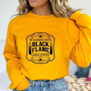 May include: Yellow sweatshirt with a black graphic print of a vintage-style label that reads "The Sanderson Sisters Black Flame Candle Company Est. 1693".