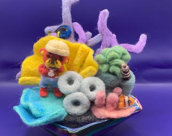 May include: A felted coral reef scene with a variety of colors. The scene includes a yellow shell, a small figure wearing a hat, and various coral formations in shades of purple, green, and pink. A small fish is also present.