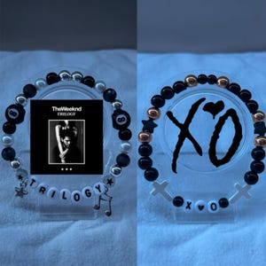 May include: Two beaded bracelets. One features black and silver beads, an 8-ball charm, and the word "TRILOGY" with a music note charm. The other has black, gold, and star-shaped beads, with "XO" and cross charms.