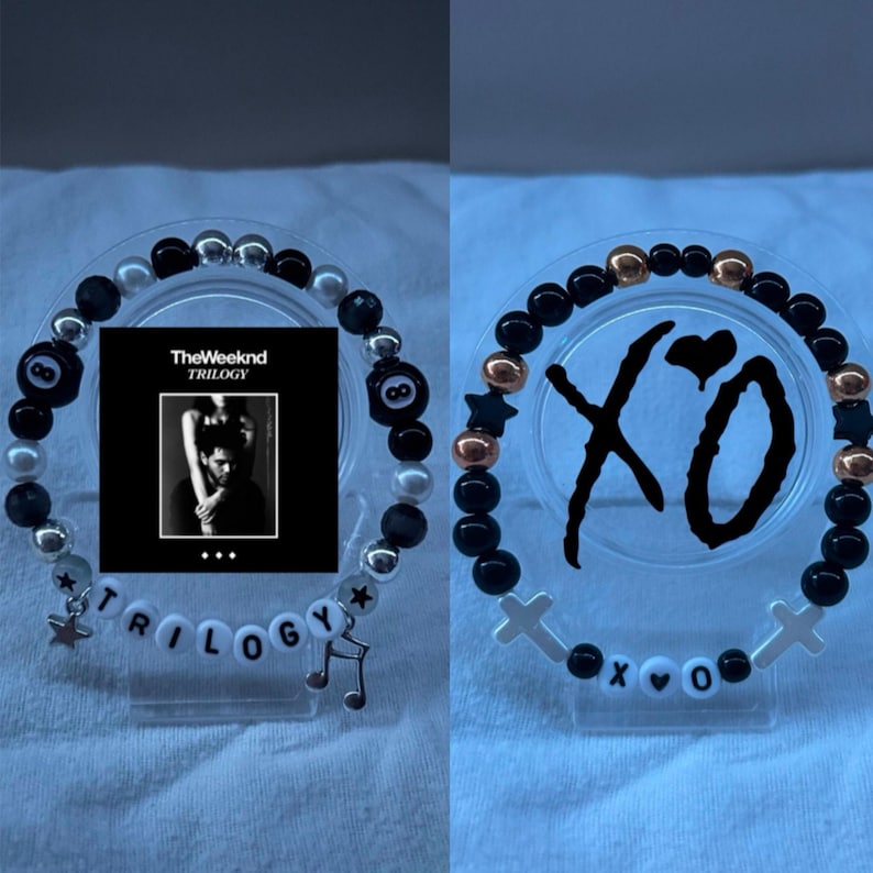 May include: Two beaded bracelets. One features black and silver beads, an 8-ball charm, and the word "TRILOGY" with a music note charm. The other has black, gold, and star-shaped beads, with "XO" and cross charms.