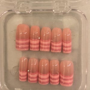 Könnte beinhalten: Ein Set künstlicher Nägel in einer transparenten Kunststoffbox. Die Nägel haben eine nackte Basis mit einem rosa gestreiften Design an den Spitzen. Einige Nägel haben einen kleinen, klaren Edelstein. Die Nägel sind in verschiedenen Größen.