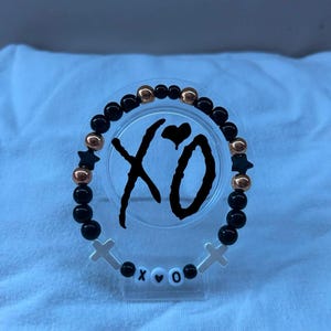 May include: A beaded bracelet with black and gold beads, star-shaped accents, and white cross charms. The bracelet features the letters "XO" and a heart symbol. The beads are strung on a clear elastic band.
