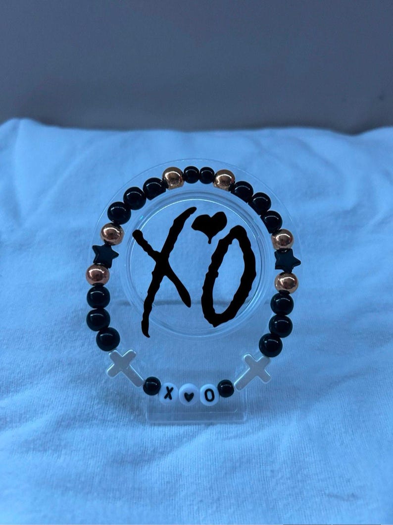 May include: A beaded bracelet with black and gold beads, star-shaped accents, and white cross charms. The bracelet features the letters "XO" and a heart symbol. The beads are strung on a clear elastic band.