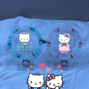 May include: Two Hello Kitty charm bracelets, one with blue beads and a boy Hello Kitty charm, the other with pink beads and a girl Hello Kitty charm. The bracelets are displayed on clear stands with a Hello Kitty charm on each.