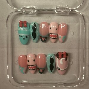 Könnte beinhalten: Ein Set aus zehn künstlichen Nägeln in einer transparenten Kunststoffbox. Die Nägel sind mit Cartoon-Gesichtern, Schnurrbärten, Schleifen und anderen Designs in Rosa, Blau, Schwarz und Weiß bemalt. Die Nägel sind oval geformt.