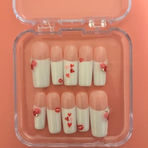 May include: A set of artificial nails in a clear plastic case. The nails feature a white base with a pink tip. Some nails have red heart and lip designs, while others have small flower embellishments.
