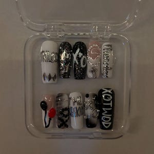 May include: A set of ten long, coffin-shaped false nails in a clear plastic case. The nails feature various designs, including text, glitter, and charms. Colors include white, black, silver, and pink. Some nails have the text "XO" and "The Weeknd".