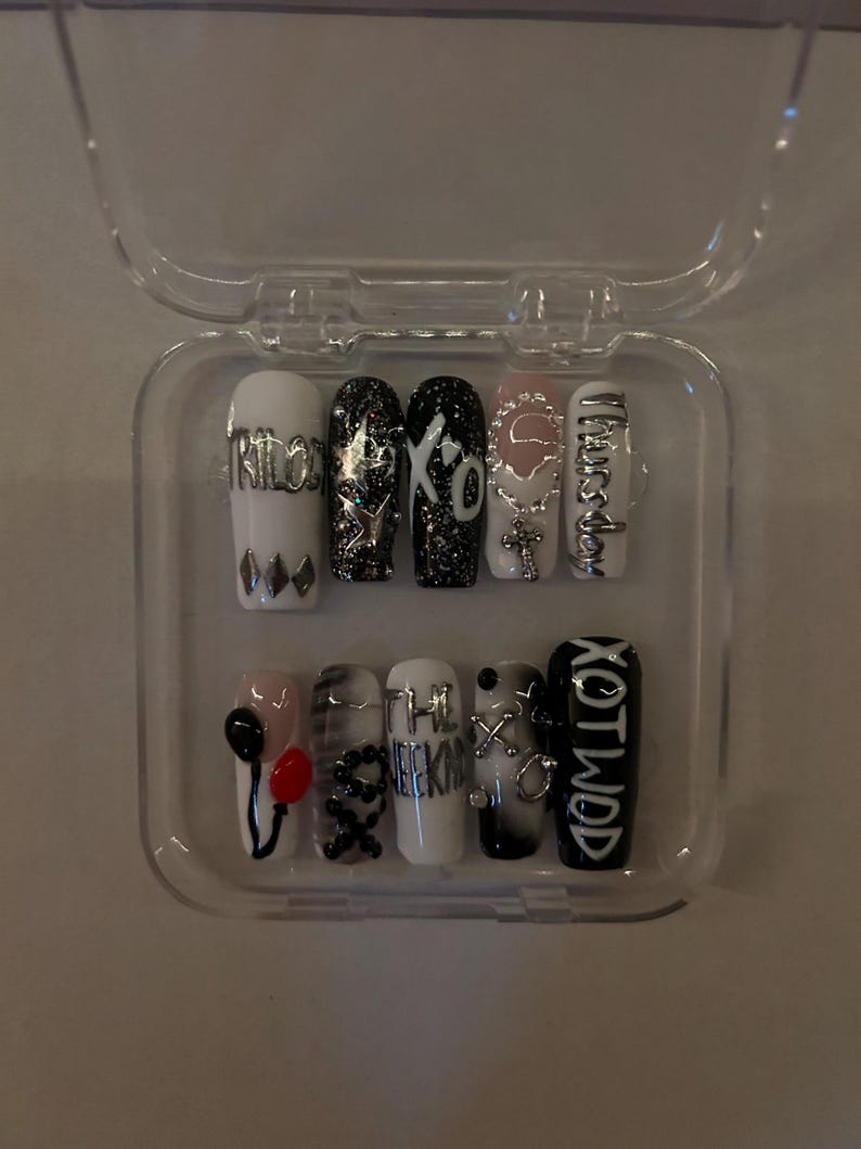 May include: A set of ten long, coffin-shaped false nails in a clear plastic case. The nails feature various designs, including text, glitter, and charms. Colors include white, black, silver, and pink. Some nails have the text "XO" and "The Weeknd".