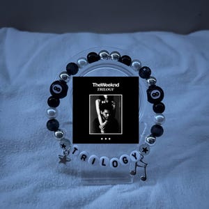 May include: A beaded bracelet featuring black, white, and silver beads. The bracelet includes a small charm of a star and a musical note. The word "TRILOGY" is spelled out with white letter beads. A small black and white image of "The Weeknd TRILOGY" is in the center.