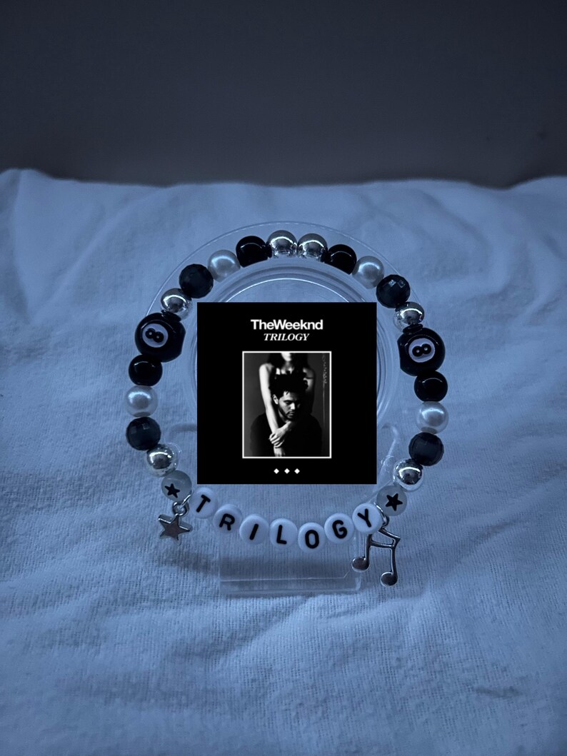 May include: A beaded bracelet featuring black, white, and silver beads. The bracelet includes a small charm of a star and a musical note. The word "TRILOGY" is spelled out with white letter beads. A small black and white image of "The Weeknd TRILOGY" is in the center.