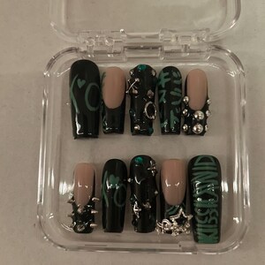 May include: A set of ten artificial nails in a clear plastic case. The nails feature a variety of designs, including black polish with green accents, nude tips, and silver embellishments. One nail reads "KISS LAND" in green lettering.
