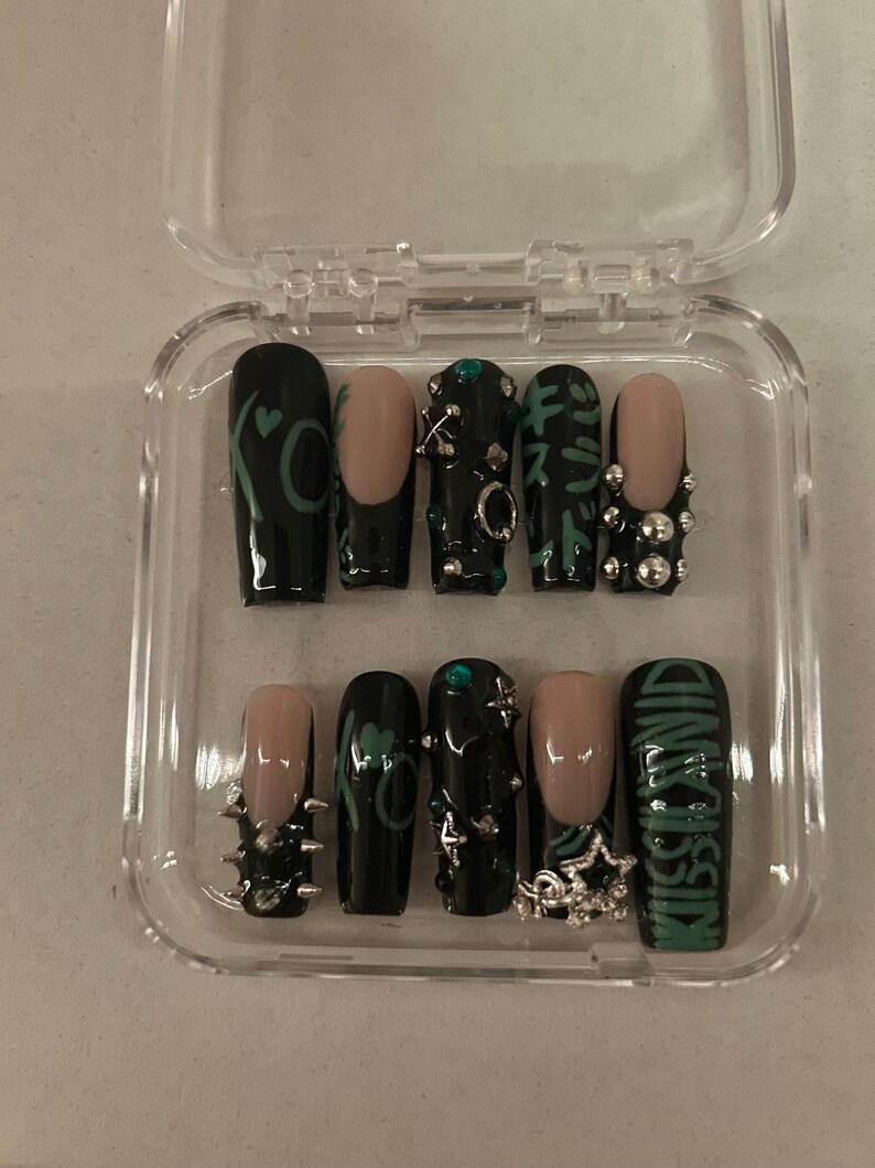 May include: A set of ten artificial nails in a clear plastic case. The nails feature a variety of designs, including black polish with green accents, nude tips, and silver embellishments. One nail reads "KISS LAND" in green lettering.