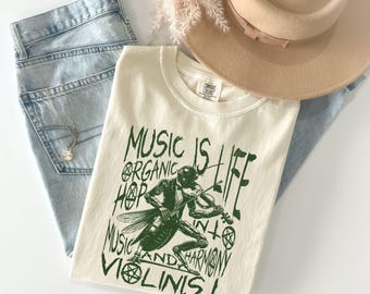 May include: Cream-colored t-shirt with a green graphic of a violin-playing insect and the words "Music is Life, Organic Hop, Music and Harmony, Violinist." The shirt is paired with light-wash jeans, a necklace, and a tan hat.
