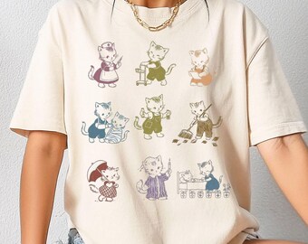 May include: Cream-colored t-shirt featuring a repeating pattern of vintage-style cat illustrations. Each cat is depicted in various poses and outfits, such as holding tools, wearing aprons, and holding an umbrella. The design has a whimsical, retro aesthetic.