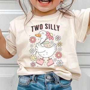 May include: Cream-colored t-shirt with the words "TWO SILLY" in pink and black. A cartoon duck wearing pink boots and holding a flower is surrounded by colorful flowers. The shirt is worn by a child.
