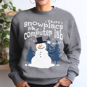 May include: A gray sweatshirt with a snowman graphic and the text "There's snowplace like computer lab." The snowman has a black top hat, a carrot nose, and black button eyes and mouth. Blue pine trees are on either side of the snowman.