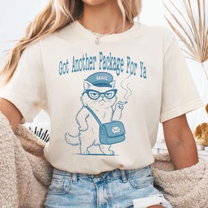May include: Cream-colored t-shirt featuring a blue illustration of a cat mail carrier wearing glasses and a hat, smoking a cigarette. The text "Got Another Package For Ya" is arched above the cat.
